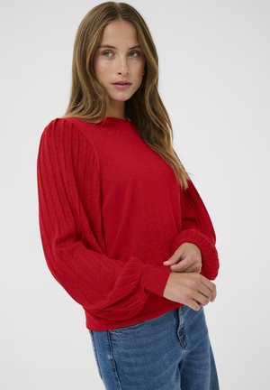 Femme aux longs cheveux bruns portant un pull long à manches longues texturé rouge et un jean bleu, posant les mains près de la taille devant un fond uni.