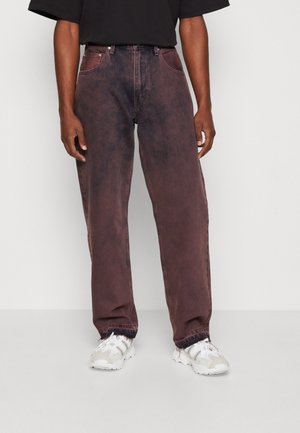 Person iført løstsiddende, mørkerødbrune vaskede jeans med rå kant kombineret med hvide og grå sneakers på lys baggrund.