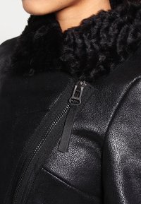 Schwarze Jacke mit einem strukturierten Kunstfellkragen, sichtbarem Reißverschluss-Detail und einer glatten, lederähnlichen Oberfläche.