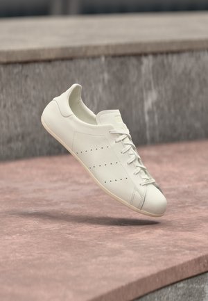 Sneaker casual bianca con lacci e piccoli dettagli traforati che vola sopra una superficie di pietra rosata e uno sfondo di pietra grigia.