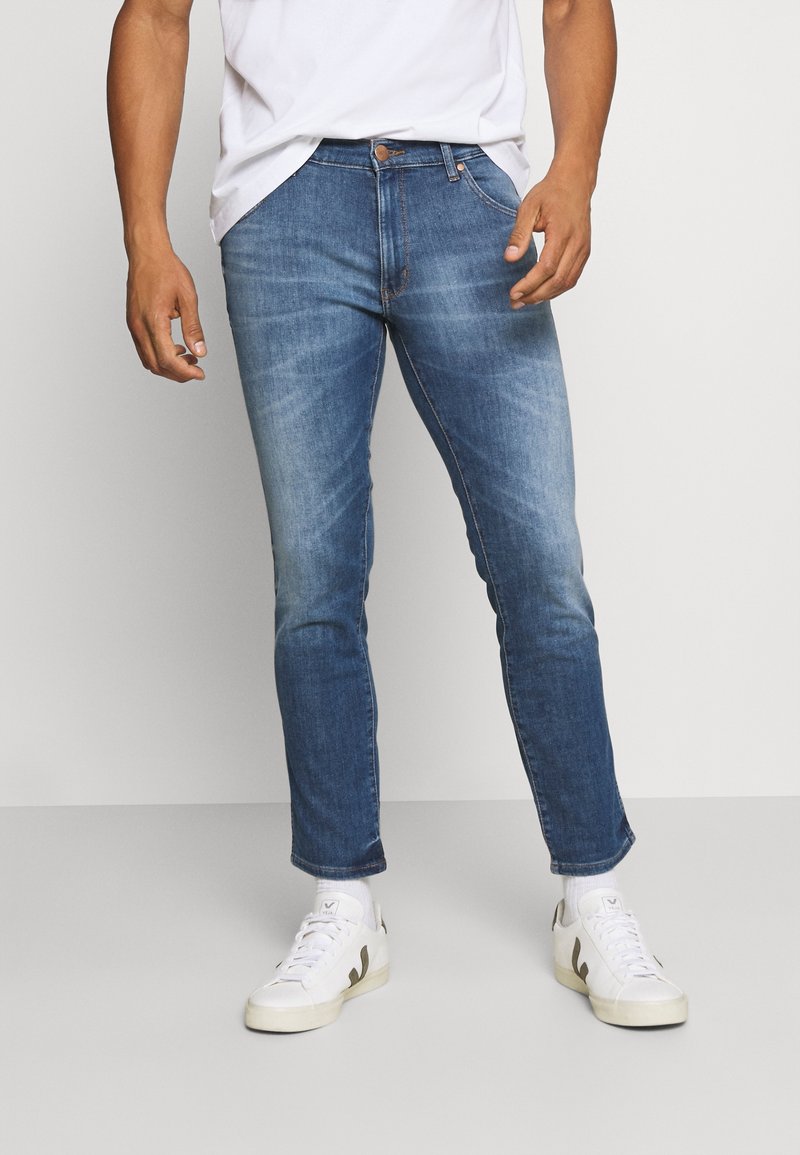 Blue denim jeans with a tapered fit, faded sections, five-pocket design, and a button closure, paired with white sneakers.