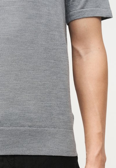 Δεξί χέρι ατόμου που φορά κοντομάνικο, ανοιχτό γκρι πλεκτό μπλουζάκι με ριμπ τελείωμα, σε απλό φόντο.