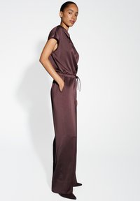 Combinaison en satin marron avec des manches courtes, une taille élastique, et des jambes larges, dotée de poches latérales et d'une texture lisse et brillante.