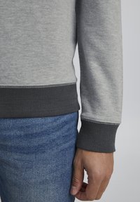 Grauer Sweatshirt mit dunkelgrauen gerippten Bündchen, aus einem strukturierten Stoff. Die Ärmel enden knapp über dem Handgelenk und sind mit blauer Denim kombiniert.