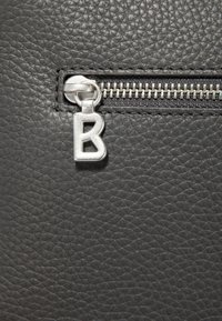 Svart texturerat läder med en dragkedjedetalj, utrustad med en silverfärgad flärp i form av bokstaven "B", som framhäver dragkedjans metalltänder och hantverk.