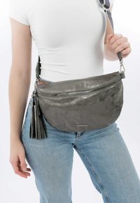 Borsa a tracolla grigia in pelle con chiusura a zip, dettaglio a frange e tracolla regolabile, indossata all'anca su un jeans in denim azzurro.