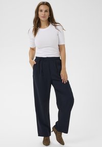 Ženska stoji z rokami v mornarsko modrih širokih hlačah, beli majici s kratkimi rokavi in leopardjih čevljih do gležnjev pred enotno podlago.