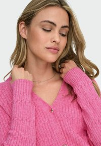 Cardigan rosa a costine con scollatura a V e dettagli di bottoni dorati. Il tessuto ha una consistenza morbida e presenta un motivo in rilievo.