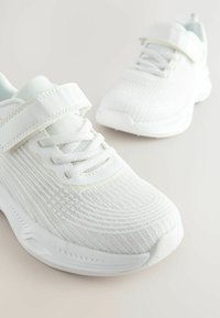 Chaussures de sport blanches avec un dessus en mesh texturé, dotées d'une sangle Velcro et de lacets, d'un bout arrondi et d'une semelle blanche rembourrée.