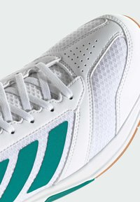 Chaussure de sport blanche avec des bandes bleu sarcelle, des lacets blancs, cuir perforé, panneaux en mesh et une semelle en caoutchouc naturel.
