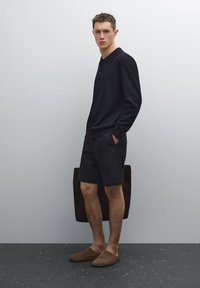 Zwarte longsleeve polo, donkere shorts en bruine instapschoenen. Model houdt een bruine tas vast. Minimalistisch ontwerp met strakke lijnen.