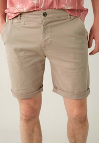 Shorts en coton beige avec un revers, dotés d'une fermeture à bouton et de poches avant, texture lisse et design épuré.