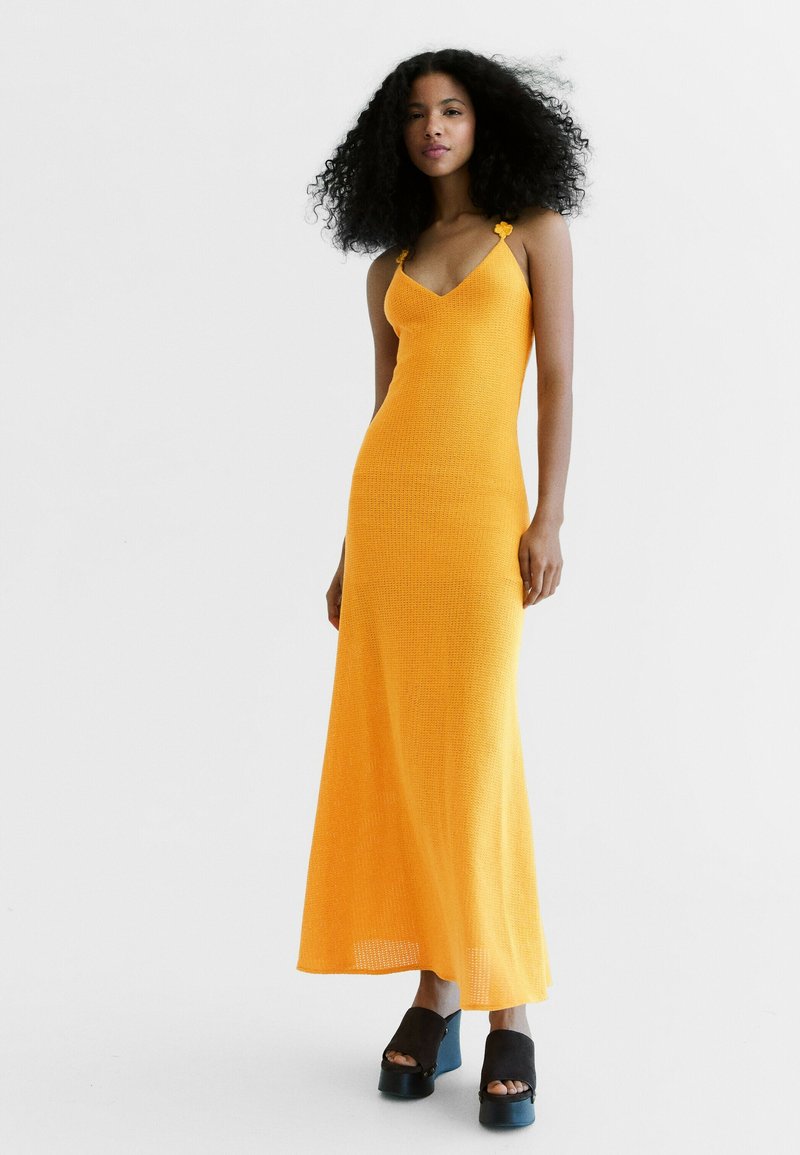 Femme aux cheveux bouclés portant une longue robe orange sans manches et des sandales compensées noires, debout devant un fond clair uni.