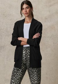 Svart ribbad cardigan med öppen front och sidofickor, i kombination med byxor med leopardmönster. Slät textur, avslappnad passform och långa ärmar.