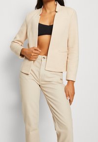 Blazer en espiga beige con un diseño sin cuello, bolsillo frontal y mangas largas, combinado con pantalones beige claros. Textura de tela suave.