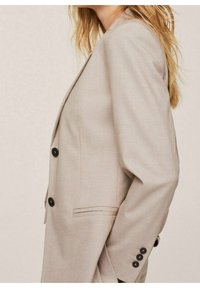 Beżowa, dopasowana marynarka na dwa czarne guziki, z wyciętymi klapami i boczną kieszenią. Gładka faktura i dopasowany krój podkreślają sylwetkę.