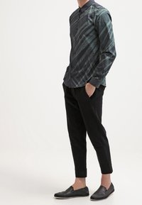 Camicia a maniche lunghe con bottoni, caratterizzata da un motivo diagonale verde scuro e nero, abbinata a pantaloni tapered neri e scarpe slip-on nere.
