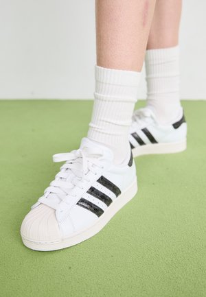 Beli usnjeni copat s črnimi lakastimi usnjenimi črtami, teksturiranim gumijastim oklepom za prste in belimi vezalkami, kombiniran z belimi rebrastimi nogavicami.