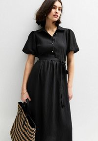 Abito nero con maniche a sbuffo corte, colletto e dettagli con bottoni. Presenta una vita cintata e un tessuto texture. Indossato con una borsa a tracolla in tessuto intrecciato.