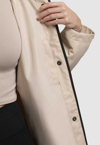 Beige jas met ritssluiting en zwarte accenten, met een soepele textuur en gestructureerd ontwerp. Zichtbare zak aan de binnenkant.