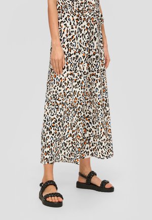 Vrouw met een lange rok met luipaardprint en zwarte sandalen met gevlochten bandjes, staand op een grijze achtergrond.