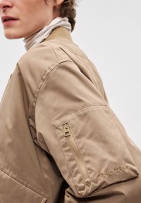 Bež bomber jakna z žepom z zadrgo na levi rokav. Ima gladko teksturo in diskretno vezenino blagovne znamke na rokavu.