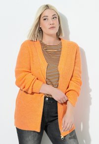Donna bionda che indossa orecchini a cerchio in argento, un cardigan di lana arancione sopra una maglietta a strisce nera e arancione, e jeans neri sbiaditi, in posa di lato.
