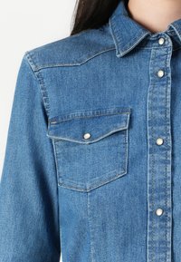 Lys blå denimskjorte med krage, knappfront og to brystlommer. Har synlige sømmer og trykknappdetaljer.