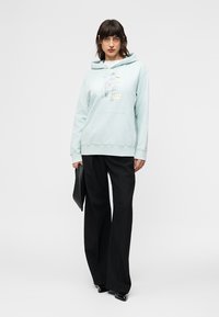 Γυναίκα που φοράει ανοιχτό μπλε φούτερ με γραφικό σχέδιο, μαύρο φαρδύ παντελόνι και μαύρα μυτερά παπούτσια, κρατώντας μαύρο φάκελο, μπροστά σε λευκό φόντο.