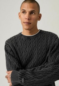 Dark gray cable-knit sweater with a round neckline and ribbed cuffs. Features a textured pattern and a small logo patch on the sleeve.