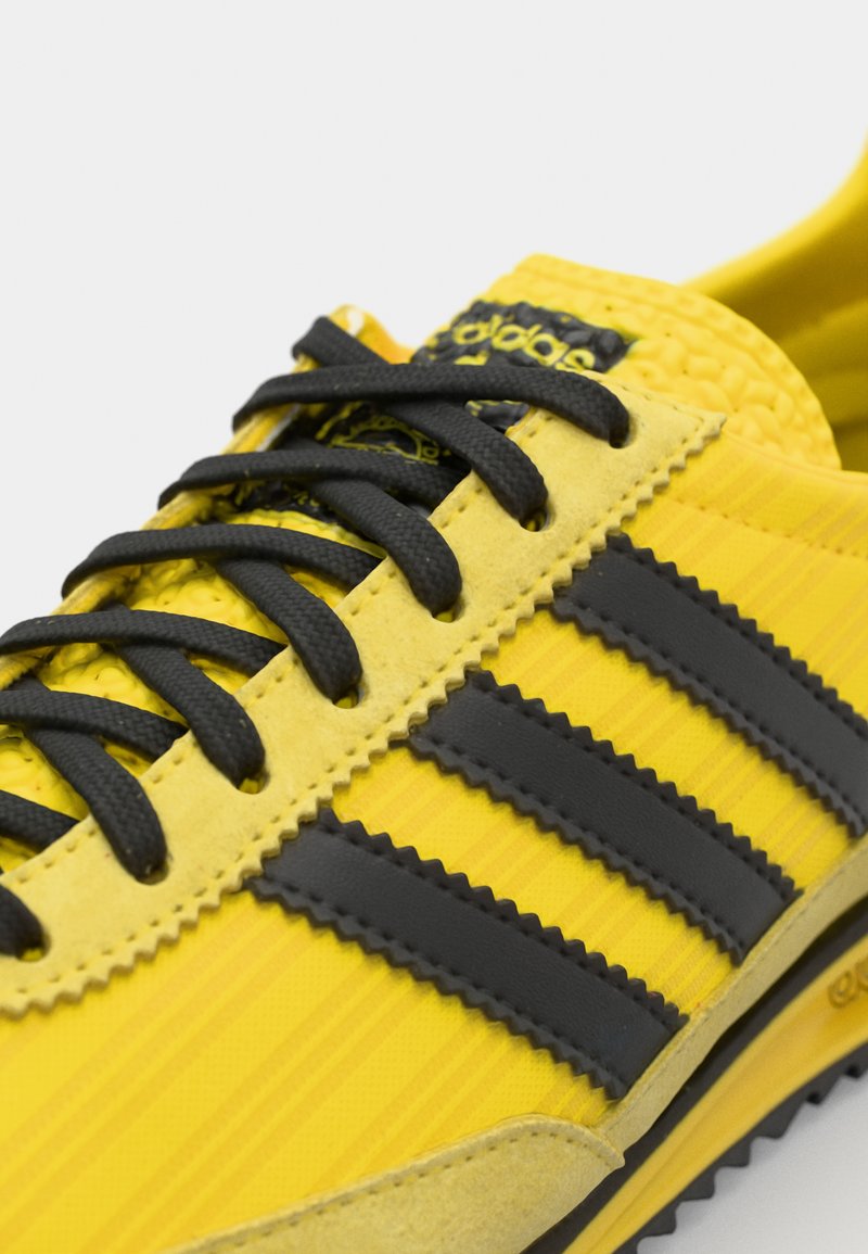 Zapatillas amarillas con cordones negros, tres rayas negras y detalles texturizados. El material está compuesto por ante y tejido sintético.