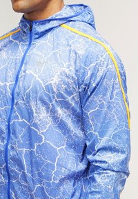 Veste bleue avec un motif craquelé texturé, dotée d'une fermeture éclair avant, d'une capuche et de rayures jaunes le long des épaules. Matière légère.