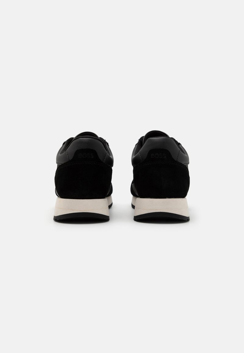 Zapatillas negras que combinan cuero liso y ante, con una entresuela blanca y una suela de goma negra, mostrando elementos de diseño minimalista.