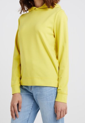 Mujer con una sudadera amarilla lisa con capucha y vaqueros azul claro, de pie contra un fondo blanco con los brazos relajados a los lados.