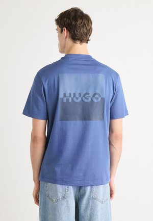 Blue cotton t-shirt featuring a large rectangular graphic on the back with the word "HUGO" in a contrasting shade. Short sleeves, round neck.