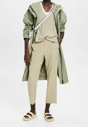 Persoon die een lichtgroene regenjas met capuchon draagt, een beige V-hals trui, cropped beige broek, witte crossbody tas en beige sandalen.