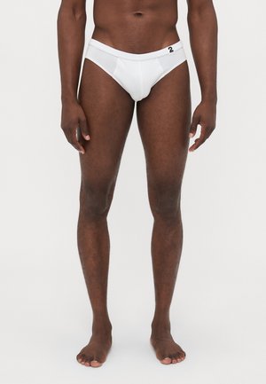 Männlicher Modell steht barfuß da und trägt weiße Unterwäsche mit einem kleinen schwarzen "2" Etikett am Bund vor einem schlichten Hintergrund.