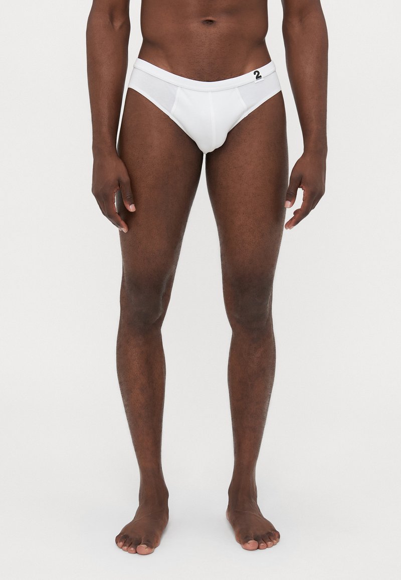 Männlicher Modell steht barfuß da und trägt weiße Unterwäsche mit einem kleinen schwarzen "2" Etikett am Bund vor einem schlichten Hintergrund.