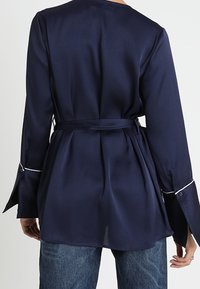Top de satén azul marino con mangas largas, cintura ceñida y detalle de ribete blanco en los puños. Textura suave, corte recto y ligera abertura en el dobladillo.