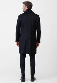 Schwarzer Wollmantel mit Kragen, knielange Ausführung, feine Struktur, Schlitz hinten, getragen über schmal geschnittenen Hosen und schwarzen Stiefeln.