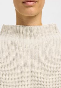 Cremefarbener gestrickter Pullover mit einem hohen Kragen, geripptem Textur und lockerer Passform, der die weiche, gewebte Struktur hervorhebt.