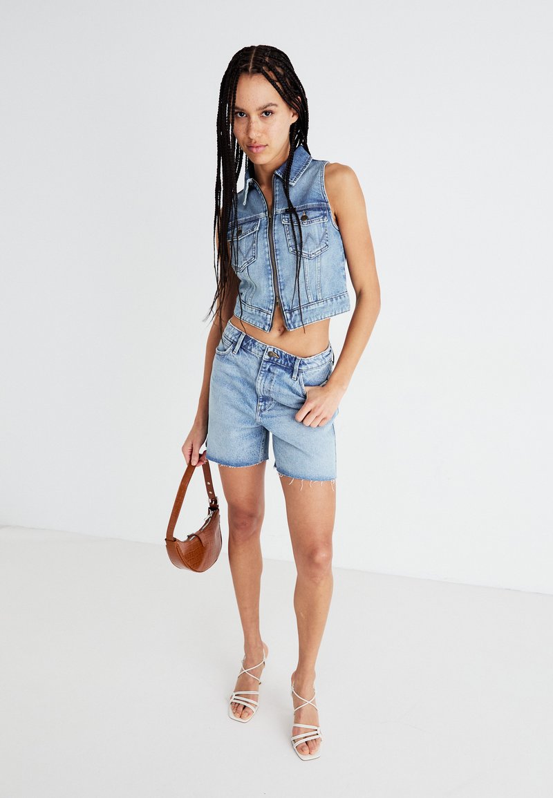 Hellblaues Denim-Crop-Top mit einem Kragen und vorderen Taschen, kombiniert mit passenden Denim-Shorts. Das Model hält eine braune Handtasche und trägt Riemchensandalen.