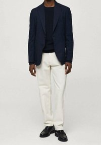 Mężczyzna ubrany w ciemnoniebieską marynarkę i sweter, jasnobeżowe spodnie oraz czarne skórzane buty, stojący na tle jasnego, jednolitego tła.