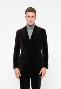 Jovem vestido com um blazer de veludo escuro de dois botões sobre um suéter de gola alta cinza, em pé frente a um fundo branco simples.