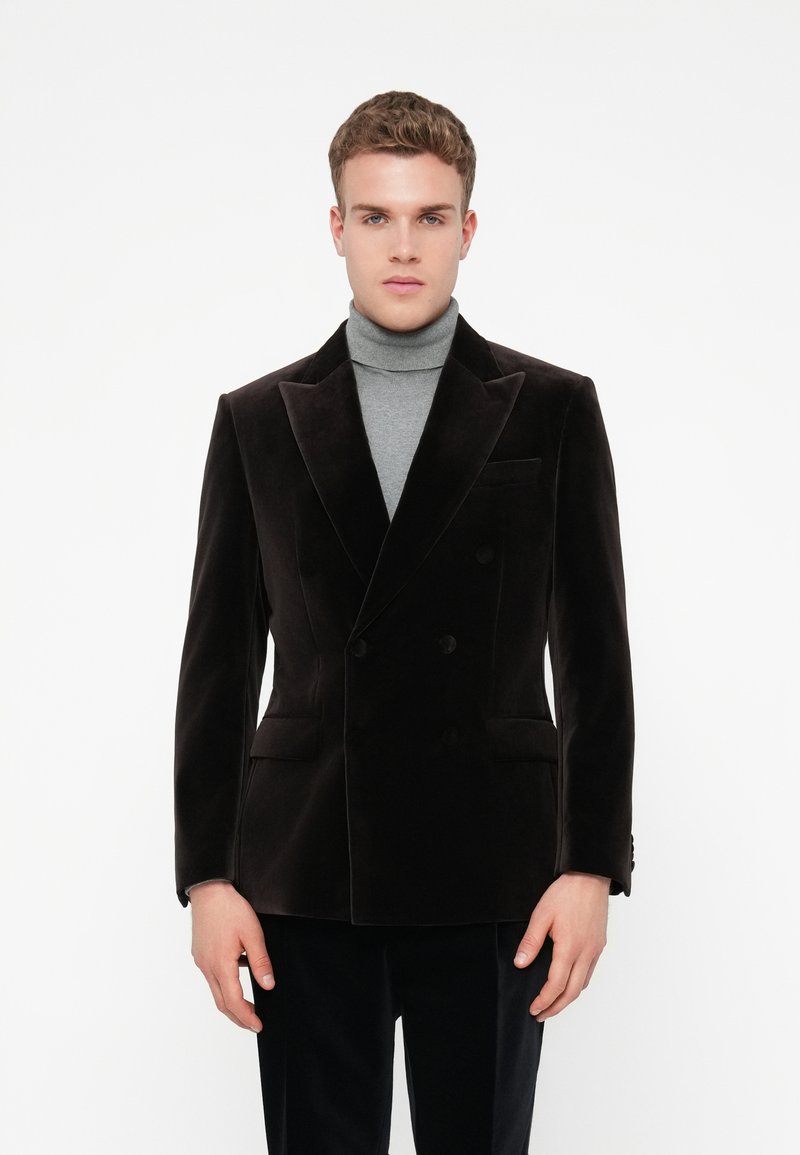 Jovem vestido com um blazer de veludo escuro de dois botões sobre um suéter de gola alta cinza, em pé frente a um fundo branco simples.