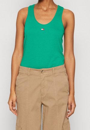 Grøn ærmeløs tank top med rund halsudskæring, med en lille rød, hvid og blå logo. Kombineret med khaki bukser. Glat stoffstruktur.