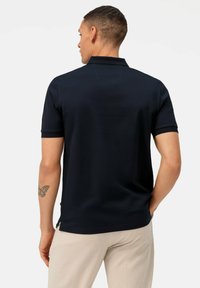 Marineblaues Polohemd mit kurzen Ärmeln, strukturiertem Stoff, klassischem Kragen und seitlichen Schlitzen. Getragen mit hellen Hosen, die ein Tattoo am Arm zeigen.