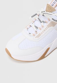 Baskets blanches en mesh et matériaux synthétiques, avec une semelle texturée, des lacets à marque et un logo bien visible sur le côté.