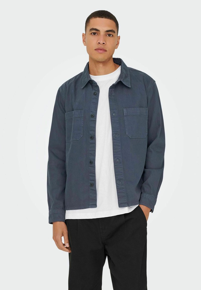 Giacca camicia in cotone blu scuro con due tasche frontali, chiusura a bottoni e colletto classico, abbinata a una t-shirt bianca e pantaloni neri.