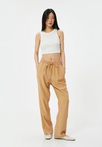 Witte, korte tanktop gecombineerd met losse, beige broek met een elastische tailleband en trekkoord, tegen een effen achtergrond.
